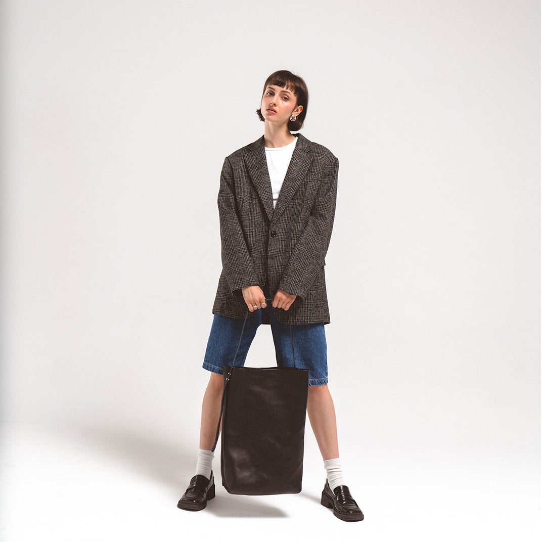 Woman holding large black leather tote bag with adjustable strap, wearing oversized blazer and denim shorts
