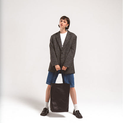 Woman holding large black leather tote bag with adjustable strap, wearing oversized blazer and denim shorts