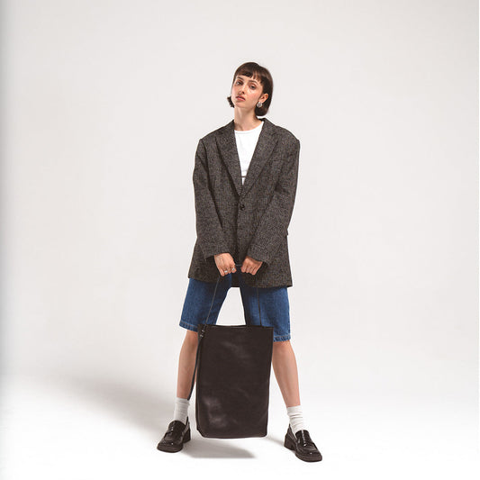 Woman holding large black leather tote bag with adjustable strap, wearing oversized blazer and denim shorts